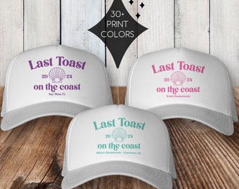 May include: Three white trucker hats with the text "Last Toast on the coast" in different colors. The hats have a mesh back and a variety of text colors including purple, pink, and teal.