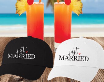 May include: A black baseball cap with white text reading "just MARRIED" and a small red heart, and a white baseball cap with black text reading "just MARRIED" and a small red heart, positioned in front of two tropical cocktails with pineapple.