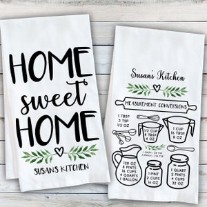 May include: Two white kitchen towels with black text and green watercolor leaves. The first towel says "Home sweet Home" and "Susan's Kitchen". The second towel says "Susan's Kitchen" and "Measurement Conversions" with a list of measurements in cups, tablespoons, teaspoons, ounces, pints, quarts, and gallons.