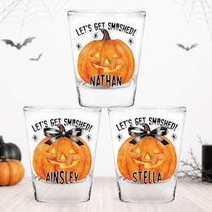 May include: Three clear glass shot glasses featuring a watercolor-style orange jack-o'-lantern pumpkin graphic. Each glass displays the text "LET'S GET SMASHED!" and a unique name: Nathan, Ainsley, or Stella. Two pumpkins feature a black bow.