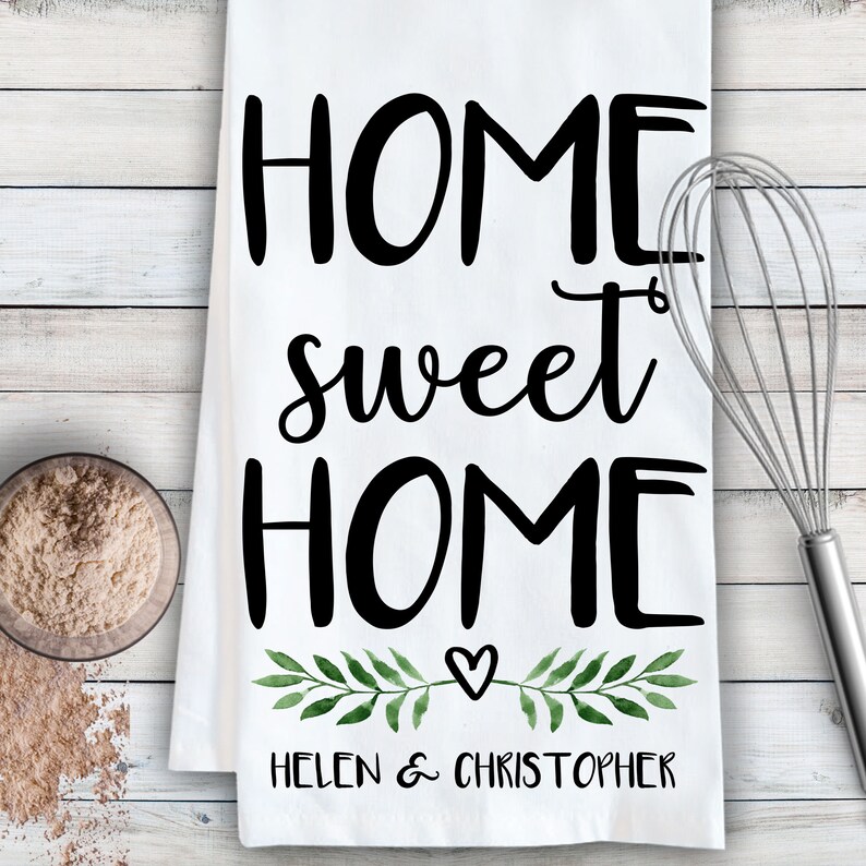 May include: White kitchen towel with the text "Home sweet home" in black cursive font. A small heart and green watercolor leaves are below the text. The text "Helen & Christopher" is at the bottom of the towel.