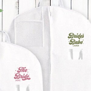 May include: Two white garment bags with a zipper closure. The bag on the left has the text "The Bride" in pink with stars on either side. The bag on the right has the text "Bride's Babe" in green with stars on either side. The name "Jackie" is printed below the text on the left bag and the name "Lauren" is printed below the text on the right bag.