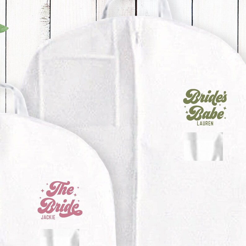 May include: Two white garment bags with a zipper closure. The bag on the left has the text "The Bride" in pink with stars on either side. The bag on the right has the text "Bride's Babe" in green with stars on either side. The name "Jackie" is printed below the text on the left bag and the name "Lauren" is printed below the text on the right bag.