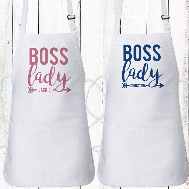 May include: Two white aprons with the text "Boss lady" in pink and blue lettering. The aprons have a pocket and adjustable straps. The name "Jackie" is printed on the pink apron and "Christina" is printed on the blue apron.