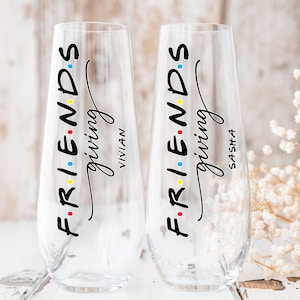 May include: Two clear stemless champagne flutes with the words "FRIENDS giving" in black script. The word "FRIENDS" has colorful dots. One glass has "Vivian" and the other has "Sasha" in black script. The glasses are on a white surface.