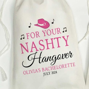 Puede incluir: Una bolsa de tela blanca con cord&oacute;n que presenta un gr&aacute;fico de sombrero de vaquero rosa con notas musicales. El texto dice: FOR YOUR NASHTY Hangover OLIVIA'S BACHELORETTE JULY 2024. El dise&ntilde;o combina letras rosas en bloque y fuente negra.