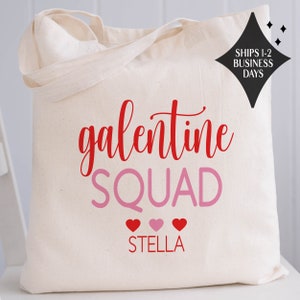May include: White canvas tote bag with the text "galentine SQUAD" in red and pink lettering. Three pink hearts are below the text, and the name "STELLA" is printed below the hearts.