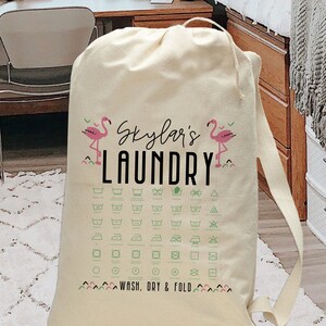 May include: A white canvas laundry bag with pink flamingos and the text "Skylar's Laundry" printed on the front. The bag has a chart of laundry care symbols and the text "Wash, Dry & Fold" at the bottom.