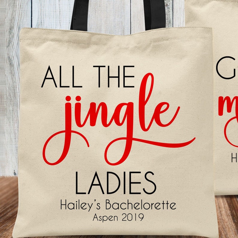 May include: A white canvas tote bag with black handles. The bag has red text that says "All the jingle ladies" and smaller black text that says "Hailey's Bachelorette Aspen 2019".