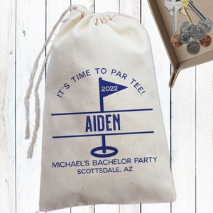 May include: A white drawstring bag with the text "It's time to par tee!" and the year "2022" printed on it. The bag also has the name "Aiden" printed on it, along with the text "Michael's Bachelor Party Scottsdale, AZ".