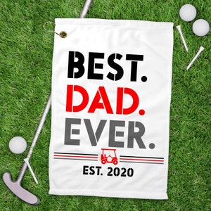 May include: White golf towel with black, red, and gray text that reads "BEST. DAD. EVER. EST. 2020". A golf cart icon is between the words "EVER" and "EST".