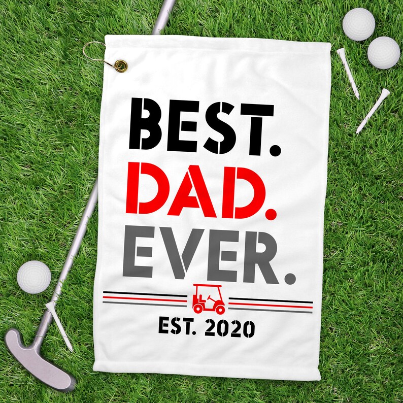 May include: White golf towel with black, red, and gray text that reads "BEST. DAD. EVER. EST. 2020". A golf cart icon is between the words "EVER" and "EST".