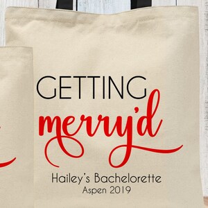 May include: A white canvas tote bag with black handles. The text "GETTING merry'd" is printed in red cursive font. Below the text is "Hailey's Bachelorette Aspen 2019" in black.