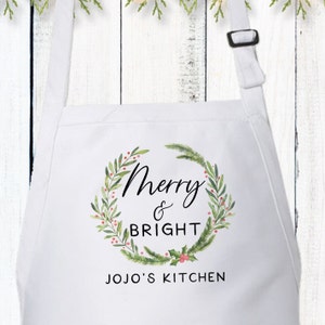 May include: White kitchen apron with a green and red watercolor wreath design. The wreath has the text "Merry & Bright" and "JOJO'S KITCHEN" printed on it.