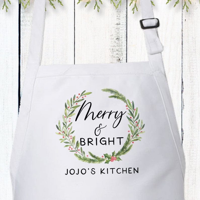 May include: White kitchen apron with a green and red watercolor wreath design. The wreath has the text "Merry & Bright" and "JOJO'S KITCHEN" printed on it.