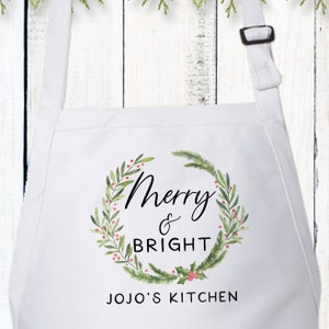 Op de afbeelding: Een wit schort met een aquarelkrans van groene dennentakken en rode bessen. In het midden staat de tekst Merry & BRIGHT in zwarte letters. Onder de krans staat de tekst JOJO'S KITCHEN in zwarte hoofdletters op het witte schort gedrukt.