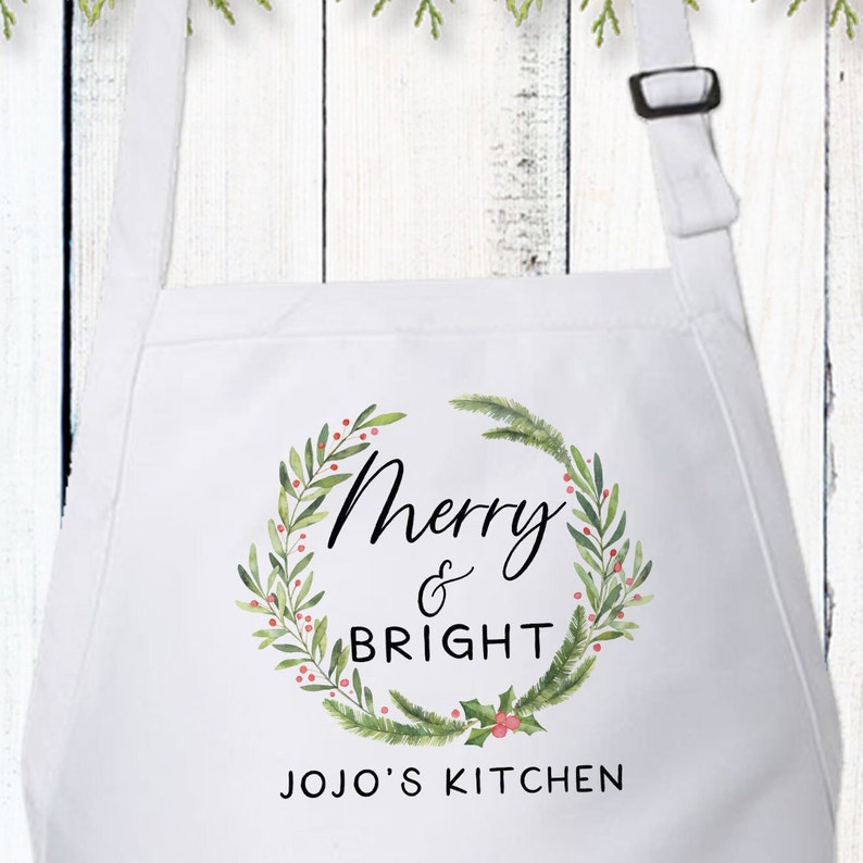 Op de afbeelding: Een wit schort met een aquarelkrans van groene dennentakken en rode bessen. In het midden staat de tekst Merry & BRIGHT in zwarte letters. Onder de krans staat de tekst JOJO'S KITCHEN in zwarte hoofdletters op het witte schort gedrukt.
