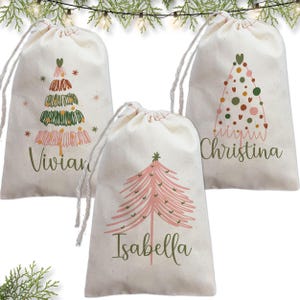 May include: Three off-white drawstring fabric gift bags featuring personalized names and Christmas tree illustrations. The left bag shows Vivian with a scribbled tree, the center shows Isabella with a pink tree, and the right shows Christina with a dotted tree.