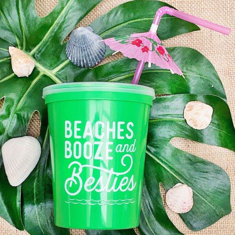 May include: Green plastic cup with the text "Beaches Booze and Besties" in white. A pink umbrella drink stirrer is in the cup. The cup is on a green leaf with seashells.