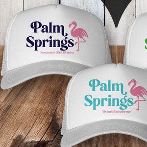 May include: Two white trucker hats with a pink flamingo and the text "Palm Springs" in blue and teal. The first hat says "Samantha's 30th Birthday" and the second hat says "Vivian's Bachelorette".