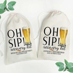 Op de afbeelding: Twee witte katoenen trekzakjes met de tekst "OH SIP! recovery kit" erop gedrukt. Eén zakje heeft de tekst "SPENCER'S BACHELOR PARTY SCOTTSDALE 2025" en het andere zakje heeft de tekst "ANTHONY'S 30TH BIRTHDAY AGED TO PERFECTION!"