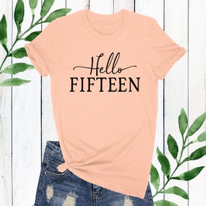 May include: Peach coloured t-shirt with black text that reads "Hello Fifteen".