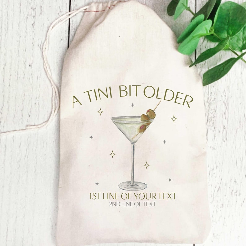 May include: A white cotton drawstring bag with the text "A TINI BIT OLDER" printed in green. A martini glass with olives is illustrated on the bag. The text "1ST LINE OF YOUR TEXT" and "2ND LINE OF TEXT" is printed below the illustration.