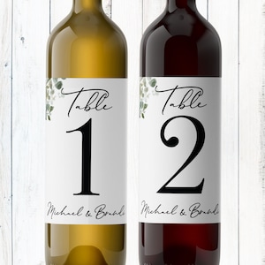 May include: Two wine bottles with labels that say "Table 1" and "Table 2" with a floral design and the text "Michael & Brandi".