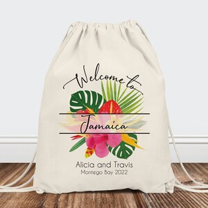 Puede incluir: Bolsa de cord&oacute;n blanco con un dise&ntilde;o floral tropical y el texto "Bienvenido a Jamaica" en escritura cursiva negra. Debajo del dise&ntilde;o floral, el texto "Alicia y Travis Montego Bay 2022".