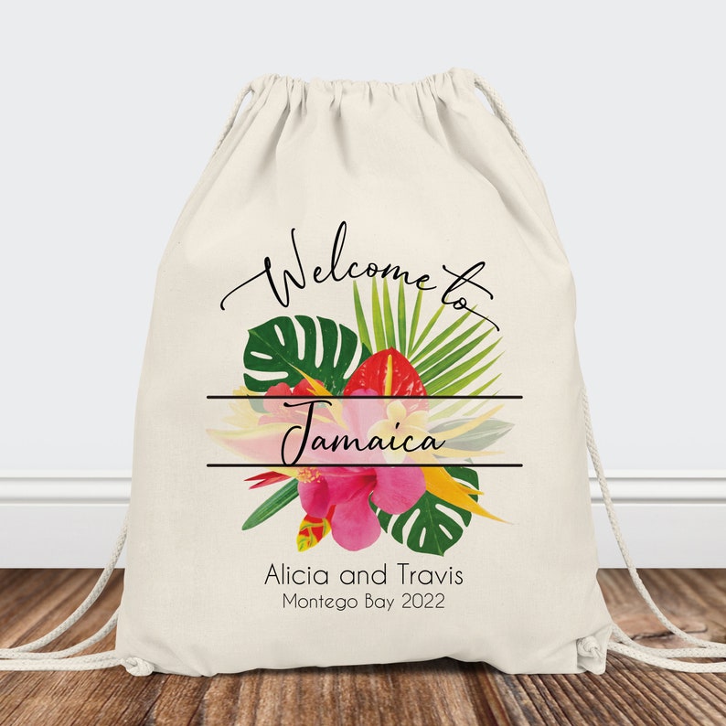 Puede incluir: Bolsa de cord&oacute;n blanco con un dise&ntilde;o floral tropical y el texto "Bienvenido a Jamaica" en escritura cursiva negra. Debajo del dise&ntilde;o floral, el texto "Alicia y Travis Montego Bay 2022".