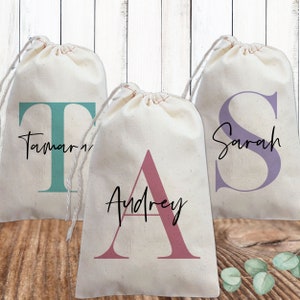 May include: Three white fabric drawstring bags with personalized names and initial letters in different colors. The bags are on a wooden surface with green leaves.