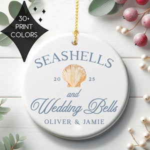 May include: A round white ceramic ornament featuring a gold hanging cord. The design includes a central seashell illustration, the text "SEASHELLS and Wedding Bells", the year "20 25", and the names "OLIVER & JAMIE" in blue lettering.