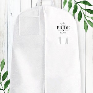Puede incluir: Bolsa de ropa blanca con cierre de cremallera. La bolsa tiene un bolsillo en la parte delantera y una etiqueta que dice "the bride - ROSE".