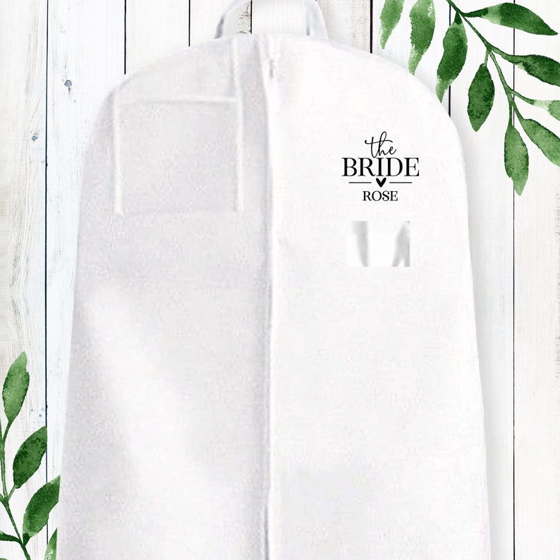 Puede incluir: Bolsa de ropa blanca con cierre de cremallera. La bolsa tiene un bolsillo en la parte delantera y una etiqueta que dice "the bride - ROSE".