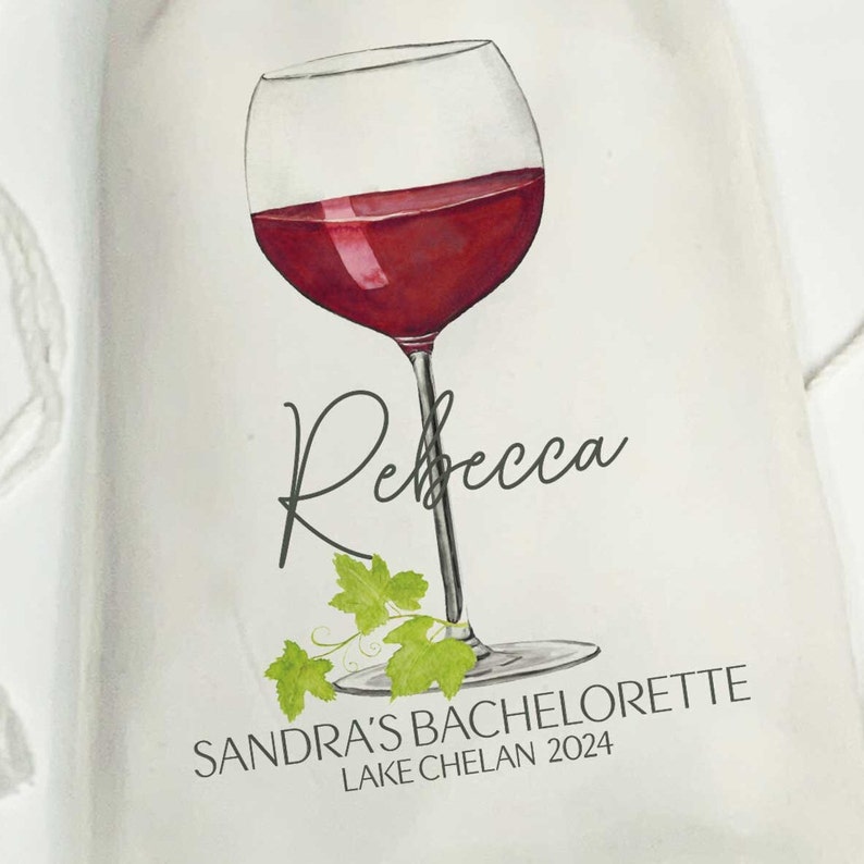 May include: A white tote bag with a watercolor illustration of a glass of red wine and a vine. The text "Rebecca" is written in cursive above the glass. The text "Sandra's Bachelorette Lake Chelan 2024" is written below the glass.