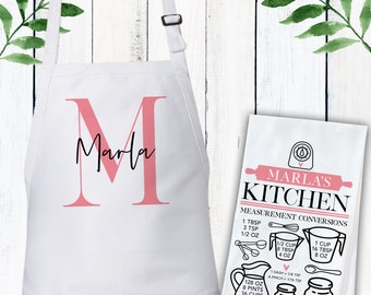 May include: A white apron featuring a large pink letter M with the name Marla in black script, alongside a white kitchen towel printed with a black and pink conversion chart labeled Marla's Kitchen, including measurements in tablespoons, cups, and ounces.