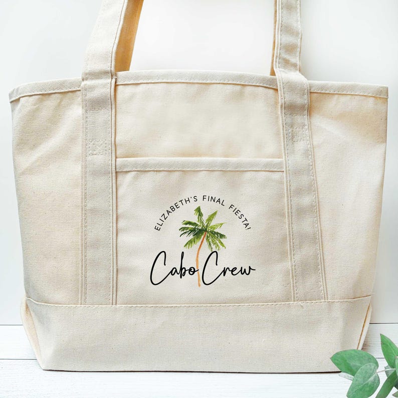 May include: A cream-colored tote bag with a front pocket and two handles. The bag features the text "ELIZABETH'S FINAL FIESTA!" around a palm tree graphic, with "Cabo Crew" written below in a script font.
