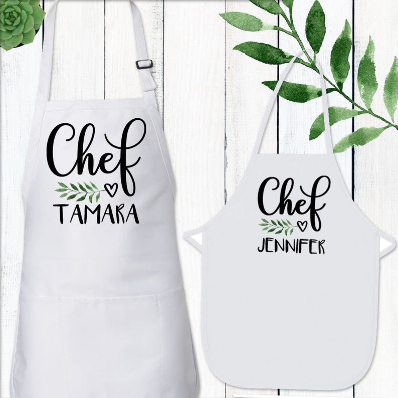 May include: Two white aprons with black text that says "Chef" with a small heart and a name below. The aprons are on a white wood background.