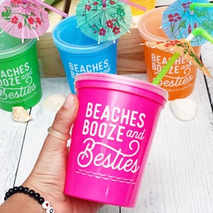 May include: Four colorful plastic cups with lids and straws. Each cup has a different color and features the text "Beaches Booze and Besties". The cups are decorated with small paper umbrellas.