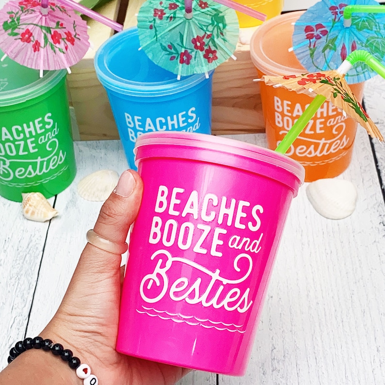 May include: Four colorful plastic cups with lids and straws. Each cup has a different color and features the text "Beaches Booze and Besties". The cups are decorated with small paper umbrellas.