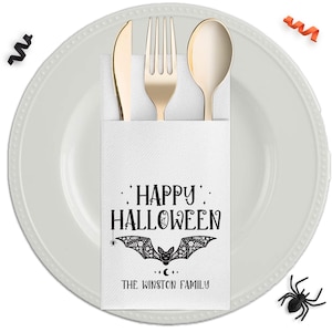 May include: A white paper napkin pocket featuring the black text HAPPY HALLOWEEN and THE WINSTON FAMILY with a decorative bat illustration, holding a gold-toned knife, fork, and spoon, placed on a white plate with black and orange confetti.