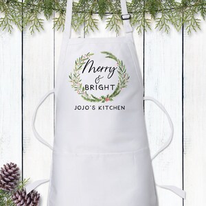 Op de afbeelding: Een wit schort met een opdruk van een groene krans met rode bessen en de zwarte tekst Merry & BRIGHT gevolgd door JOJO'S KITCHEN. Het schort is voorzien van een verstelbare nekband met een zwarte gesp en twee voorzakken voor extra gemak.