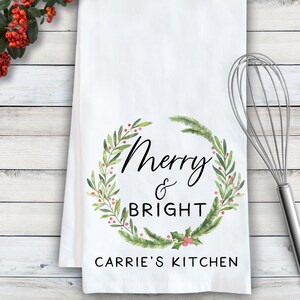 May include: White kitchen towel with a green and red watercolor wreath design. The wreath has the text "Merry & Bright" in black lettering. The bottom of the towel has the text "Carrie's Kitchen" in black lettering.
