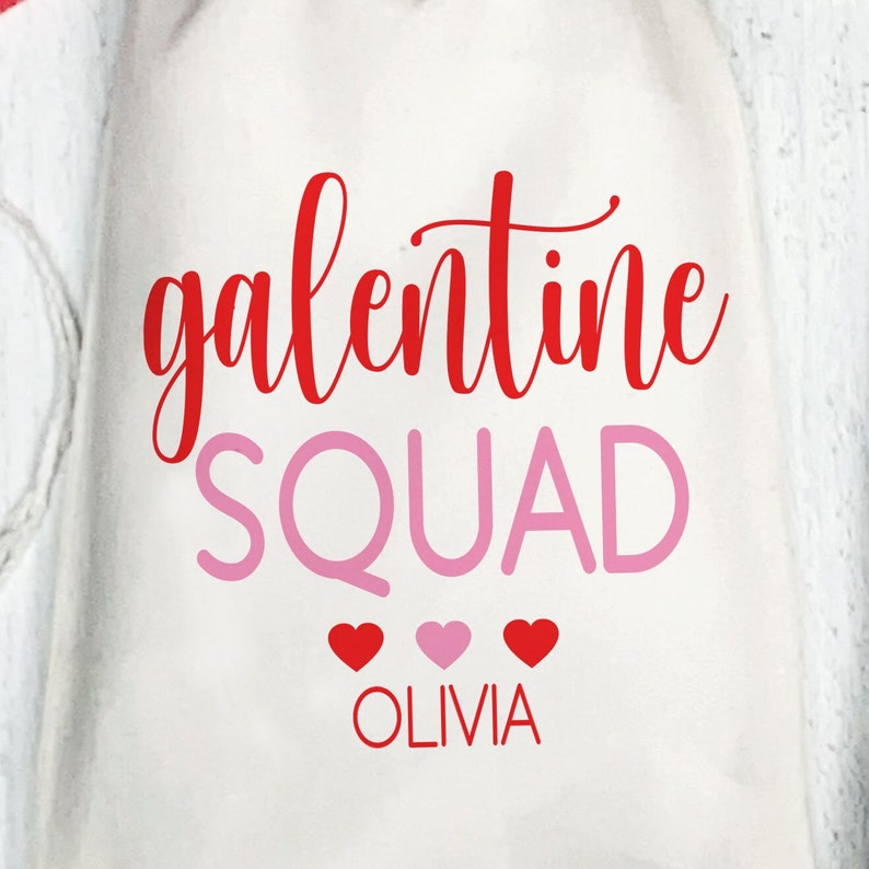 May include: White canvas tote bag with the text "galentine squad" in red cursive lettering and "SQUAD" in pink block lettering. Three red hearts and one pink heart are below the text, and the name "OLIVIA" is written in red below the hearts.