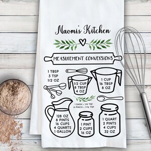 May include: A white kitchen towel with black text and illustrations of kitchen tools and measuring cups. The text reads "Naomi's Kitchen" and "Measurement Conversions". The towel includes conversion charts for ounces, cups, pints, quarts, and gallons.