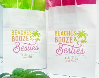 May include: Two white paper gift bags with handles. Both bags have a graphic design that says "Beaches Booze & Besties" in yellow, pink, and blue lettering with palm trees. The text "La Jolla, CA April 2023" is printed on the bottom of each bag.
