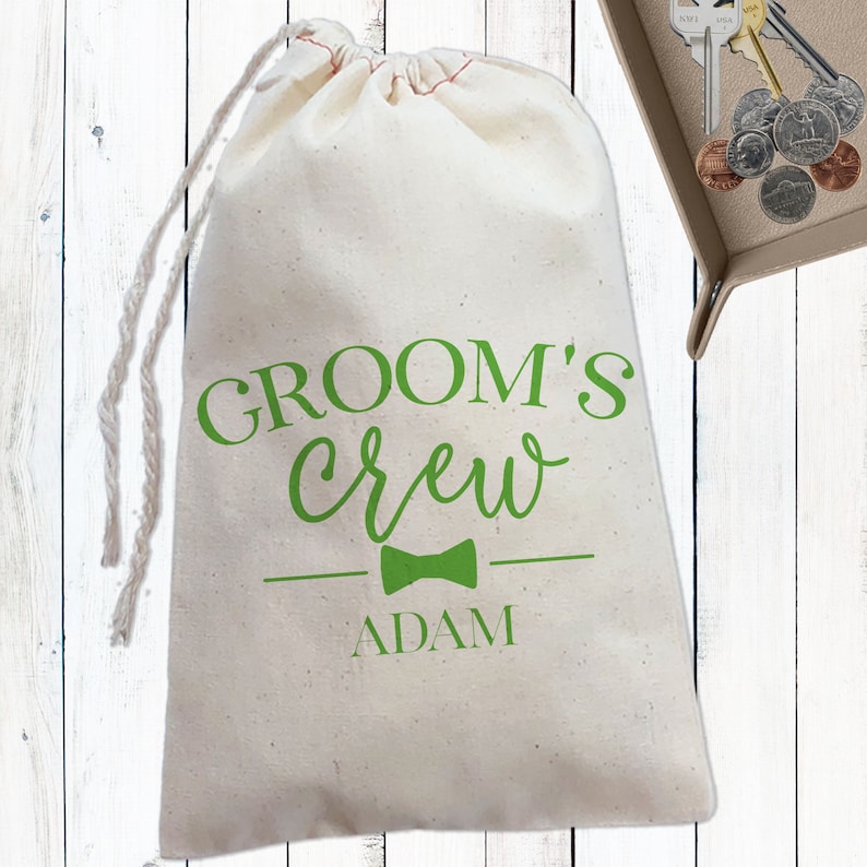 May include: A white drawstring bag with the text "Groom's Crew" in green lettering, a green bow tie, and the name "Adam" printed below.