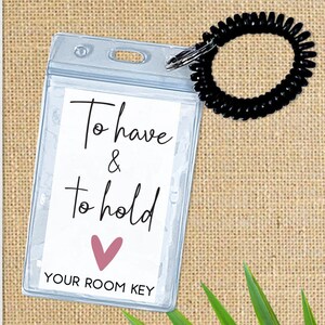 May include: A clear plastic badge holder with a black spring coil keychain. The badge holder has a white card inside with the text "To have & to hold" in black cursive writing and a pink heart. The card also has the text "YOUR ROOM KEY" in black.