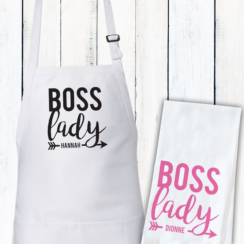 May include: White kitchen apron with black text that reads "Boss lady" with an arrow and the name "HANNAH". A white tea towel with pink text that reads "Boss lady" with an arrow and the name "DIONNE".