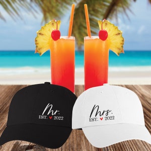 May include: Two black and white baseball caps with the words "Mr. EST. 2022" and "Mrs. EST. 2022" embroidered on them. The caps are sitting on a wooden surface with two tropical drinks behind them.